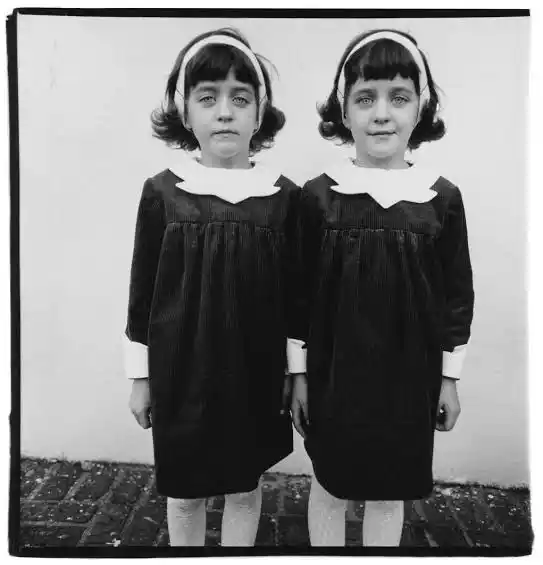 Dos niñas gemelas posan de frente con vestidos oscuros y cuellos blancos en una fotografía de Diane Arbus